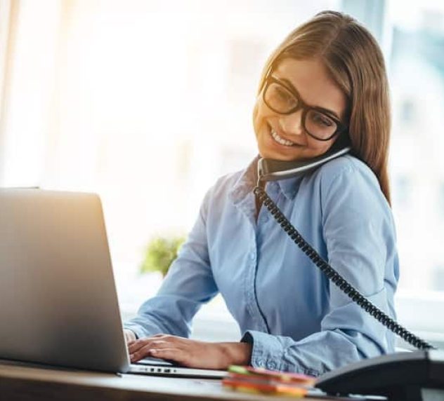 Cheerful young beautiful woman in glasses talking on the phone and using laptop with smile while sitting at her working place